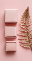 Fototapeta premium Three pale pink velvet boxes vertically aligned beside a fern frond on a matching pink background; minimalist aesthetic