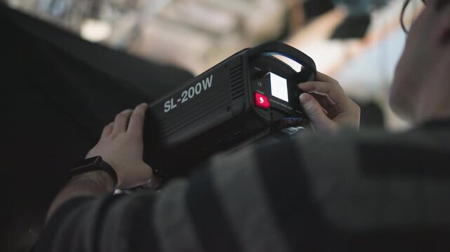 camera technician adjusting brightness knob and power switch on SL-200W LED studio light mounted on stand, manipulating control panel with cables and blue backdrop during broadcast production