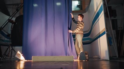 Young male stagehand pulls blue curtain during television studio setup, floor spotlight casts shadow on striped backdrop while crane camera gear and cables visible backstage broadcast production