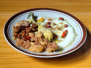 A dish of chicken porridge with yellow sauce, chicken pieces, omelet, orek tempeh and fried peanuts, served on a brown enamel plate. Chicken porridge, traditional Indonesian food.