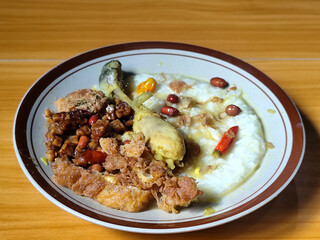 A dish of chicken porridge with yellow sauce, chicken pieces, omelet, orek tempeh and fried peanuts, served on a brown enamel plate. Chicken porridge, traditional Indonesian food.
