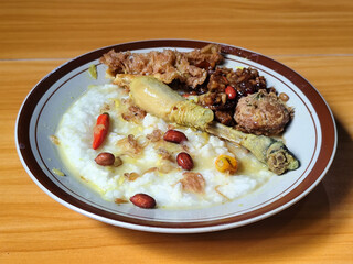 A dish of chicken porridge with yellow sauce, chicken pieces, omelet, orek tempeh and fried peanuts, served on a brown enamel plate. Chicken porridge, traditional Indonesian food.