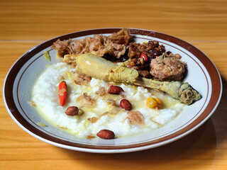 A dish of chicken porridge with yellow sauce, chicken pieces, omelet, orek tempeh and fried peanuts, served on a brown enamel plate. Chicken porridge, traditional Indonesian food.