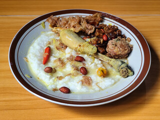 A dish of chicken porridge with yellow sauce, chicken pieces, omelet, orek tempeh and fried peanuts, served on a brown enamel plate. Chicken porridge, traditional Indonesian food.