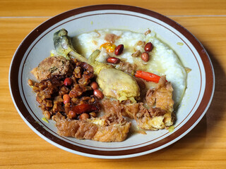 A dish of chicken porridge with yellow sauce, chicken pieces, omelet, orek tempeh and fried peanuts, served on a brown enamel plate. Chicken porridge, traditional Indonesian food.