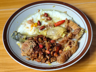 A dish of chicken porridge with yellow sauce, chicken pieces, omelet, orek tempeh and fried peanuts, served on a brown enamel plate. Chicken porridge, traditional Indonesian food.