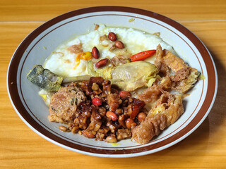 A dish of chicken porridge with yellow sauce, chicken pieces, omelet, orek tempeh and fried peanuts, served on a brown enamel plate. Chicken porridge, traditional Indonesian food.