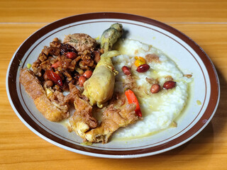 A dish of chicken porridge with yellow sauce, chicken pieces, omelet, orek tempeh and fried peanuts, served on a brown enamel plate. Chicken porridge, traditional Indonesian food.