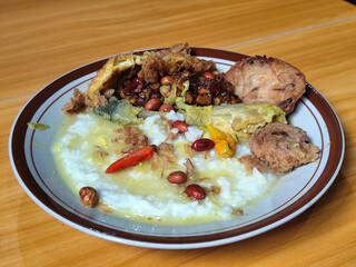 A dish of chicken porridge with yellow sauce, chicken pieces, omelet, orek tempeh and fried peanuts, served on a brown enamel plate. Chicken porridge, traditional Indonesian food.