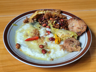 A dish of chicken porridge with yellow sauce, chicken pieces, omelet, orek tempeh and fried peanuts, served on a brown enamel plate. Chicken porridge, traditional Indonesian food.