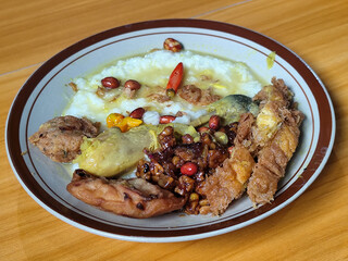 A dish of chicken porridge with yellow sauce, chicken pieces, omelet, orek tempeh and fried peanuts, served on a brown enamel plate. Chicken porridge, traditional Indonesian food.