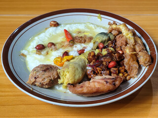 A dish of chicken porridge with yellow sauce, chicken pieces, omelet, orek tempeh and fried peanuts, served on a brown enamel plate. Chicken porridge, traditional Indonesian food.