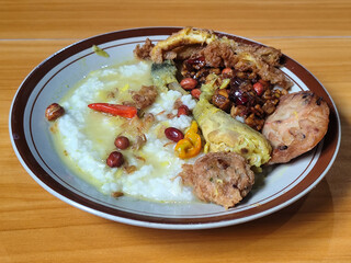 A dish of chicken porridge with yellow sauce, chicken pieces, omelet, orek tempeh and fried peanuts, served on a brown enamel plate. Chicken porridge, traditional Indonesian food.