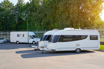Two modern caravans are parked side by side in a sunlit area. Lush green trees surround the lot,...