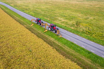 Drone view of two tractors with a mechanical mowers mowing grass on the side of the asphalt road