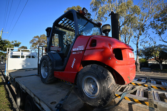 Rear corner of a red and black Manitou MH25-4 forklift truck, sitting atop a trailer, during a sunny day