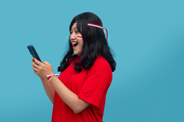 Naklejka premium Smiling indonesian woman looks at phone while holding flag. Ideal for cheerful notification, social media post, or Indonesia Independence Day concept