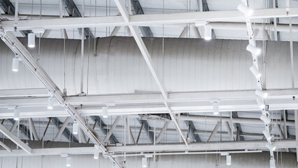 White LED spotlight lamps hanging on the ceiling frame under steel roof truss structure with air duct pipe system nearby. Lighting design installation in modern warehouse or wide span building