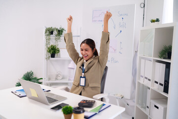 Asian adult businesswoman celebrates major success achieving profit targets with triumphant joy at modern office workplace desk professional growth career achievement