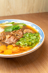 Asian beef noodle soup served in broth with fresh bok choy, daikon, carrots, and cilantro in a ceramic bowl on wooden table.