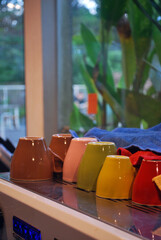 Colorful espresso cups drying upside down on a coffee machine, partially covered with microfiber cloths, with green plants in the background. Cozy café or barista workspace vibe.