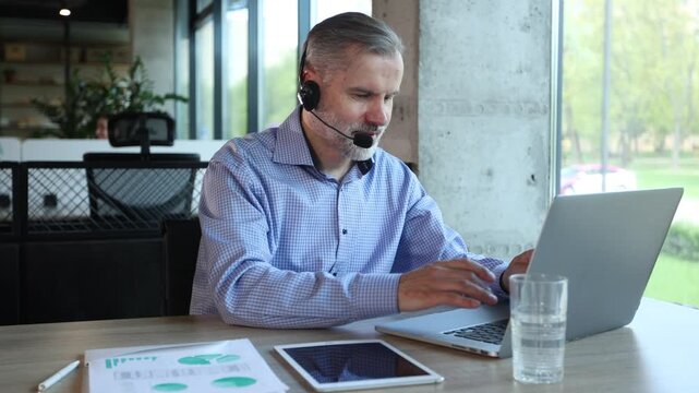Handsome gray-haired man wearing a headset, sitting in call-center office, smiling and working