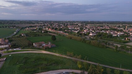 Fototapeta premium Aerial view of Castelvetro Piacentino with green fields, rural homes, and distant townscape at dusk