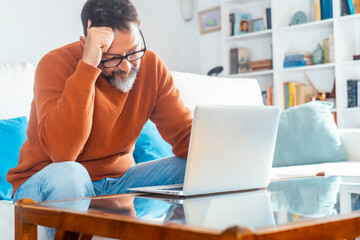 Middle-aged man working from home on laptop in living room, sitting on sofa with table, focused and stressed freelancer dealing with bills, household economy, deadlines and responsibilities