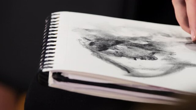 Student uses piece of charcoal to add dark tones to drawing in spiral-bound sketchbook. Artwork skretch in process with areas of shading
