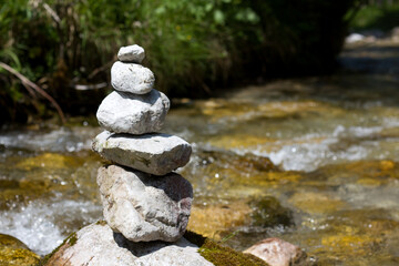 A cairn of five rocks stands by a flowing stream, with green foliage in the blurred background under natural light
