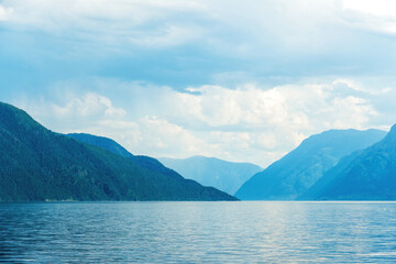 A calm lake reflects the towering, forested mountains under a cloudy sky. The cool tones and gentle ripples create a peaceful landscape