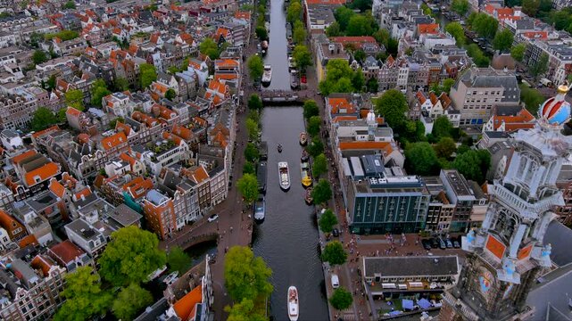 4K aerial over Amsterdam&rsquo;s Prinsengracht with the Westerkerk tower, boats beneath canal bridges, and rows of Dutch gabled houses - vivid UNESCO heritage, tourism and everyday urban life.