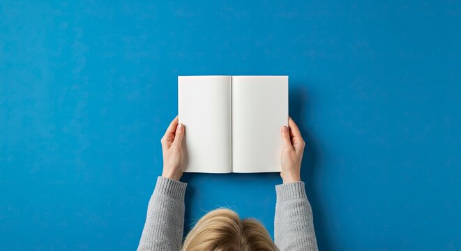 A person holds open a blank notebook against a solid blue background viewed from directly overhead