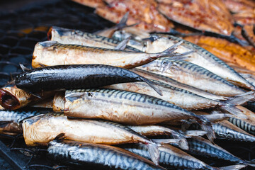 Freshly smoked mackerel fish stacked on a grill at an outdoor market. The fish display their distinctive silvery skin and rich texture, ready for sale or consumption