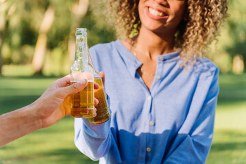 Friends toasting beer bottles, laughing together under bright sunlight in verdant park, sharing carefree summer moment