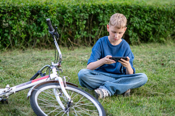 Fototapeta premium Blond teenage boy playing on smartphone while sitting on grass next to bicycle