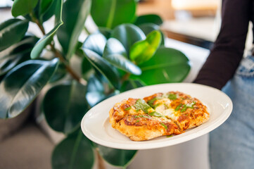 Homemade pizza with cheese and herbs on a white plate held by hand, against a lush green indoor plant background. Bright, fresh and appetizing scene.