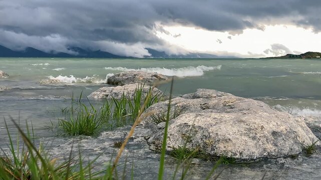 Large rocks and plants line windswept lake shore, dramatic clouds, storm waves