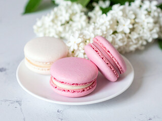 Fototapeta premium A plate with three macarons of different pink and white colors. Behind the plate is a cluster of white lilac flowers.