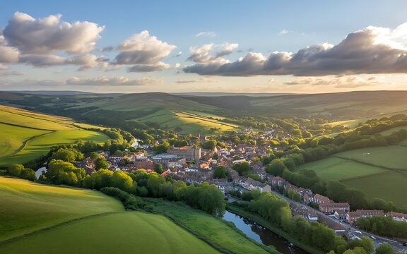 Aerial view of a picturesque English village nestled in rolling green hills at sunset with dramatic clouds Keywords: aerial, village, town, countryside, England, UK, British, rural, landscape