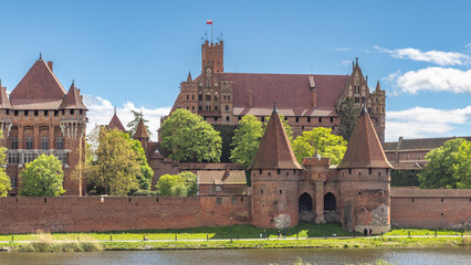 Naklejka premium Malbork Castle, a brick Gothic castle in northern Poland, Europe. Magnificent medieval castle, a testament to history and architecture, stands proudly against a backdrop of blue sky and lush greenery.
