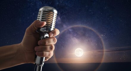 A hand holding a classic microphone against a starry night sky with a glowing full moon