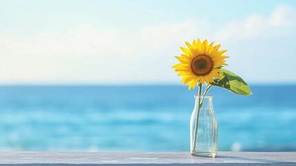 A single sunflower in a clear glass vase placed on a concrete surface, with a calm blue ocean in the background. 