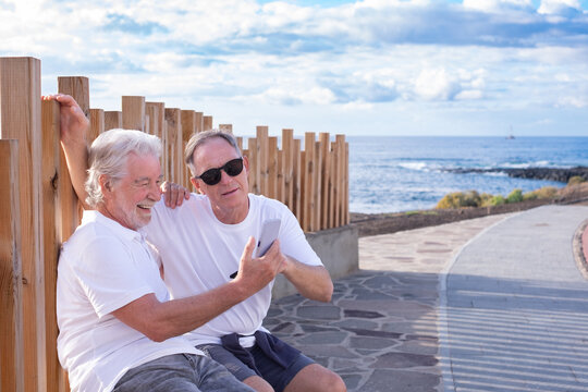 Senior couple of men sitting outdoors in the sunset light looking at mobile phone, two carefree pensioners together by the sea enjoying tech and social apps - Powered by Adobe