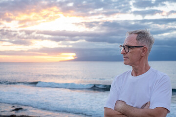 Fototapeta premium Senior mature man with crossed arms at the sea beach at sunset light looking the horizon over sea appreciating freedom and nature, handsome man enjoys a relaxed retirement lifestyle