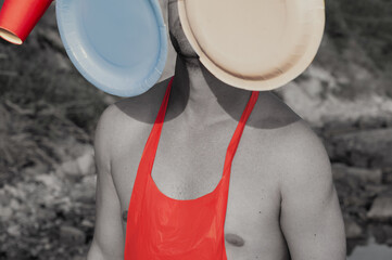 Conceptual surreal portrait of a shirtless man in a red plastic apron, his face obscured by floating disposable plates and a red cup. 