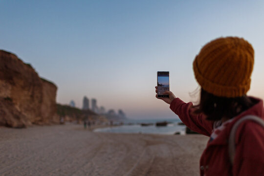Close up of female hands holding smartphone and taking photos of amazing orange city sunset on beach. Beautiful panoramic view of sunset. social media content.
