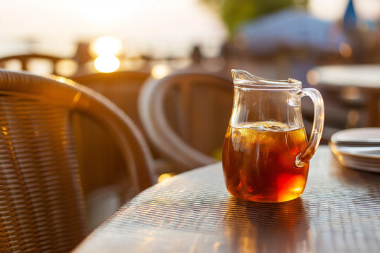 Pitcher of iced tea on outdoor table with woven chairs, amber sunlight passing through liquid, calm summer evening