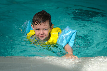Small boy learning to swim in a swimming pool on a sunny day. Happy kid playing in clear water with armbands. Childhood summer vacation and water safety concept. A child aged four years.