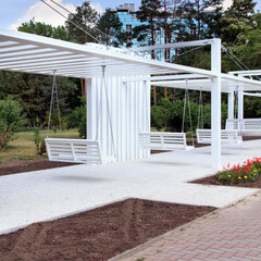 A modern outdoor space featuring white swings under a minimalist structure. The area is surrounded by greenery and flower beds